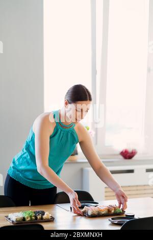 Young woman waiting for guests, friends while arranging the table with various sushi sets in a sunny home with window in the background Stock Photo