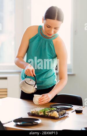 Smiling woman pouring tea while waiting her friends for a sushi party in a sunny living room Stock Photo