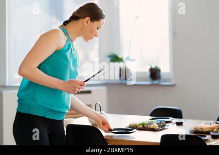 Young woman waiting for guests, friends while arranging the table with various sushi sets in a sunny home with window in the background Stock Photo