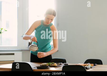 Smiling woman pouring tea while waiting her friends for a sushi party in a sunny living room Stock Photo