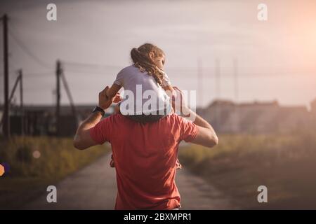 Dad dancing on his shoulders with his daughter in sun. Father travels with baby on his shoulders in rays of sunset. Child with parents walks at sunset Stock Photo