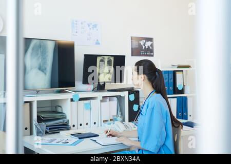 Side view portrait of young female medic looking at chest x-ray images and writing on clipboard while sitting at desk in clinic, copy space Stock Photo