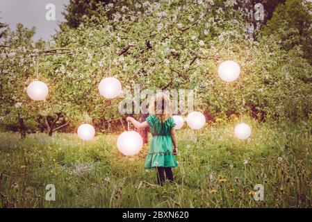 Cute blonde curly hair girl standing in home apple garden, holding glowing round paper lantern and lot of lanterns hanging from blooming apple tree. E Stock Photo