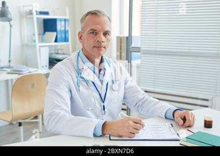 Portrait of handsome mature doctor wearing lab coat and looking at camera while sitting at desk in office of modern clinic, copy space Stock Photo