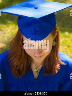 Young Adult Red Haired Pale Faced Female in a Blue Graduation Cap and Gown taken from Above with Green Grass in the Background Stock Photo