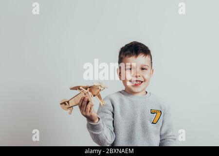 Photo of an adorable boy who looks into the camera, smiles and plays with a handmade wooden plane on a gray background. Vintage tone Stock Photo
