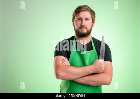 Supermarket employee with green apron and black t-shirt, he confidently holds a knife in his hand isolated on green background Stock Photo