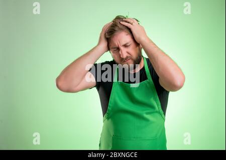 Supermarket employee with green apron and black t-shirt, has headache isolated on green background Stock Photo