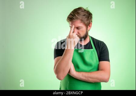 Supermarket employee with green apron and black t-shirt, wondering isolated on green background Stock Photo