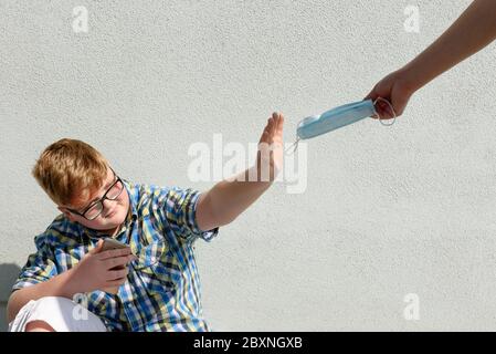The red-haired boy with glasses refuses to receive the surgical mask offered by an adult. Boy with glasses and plaid shirt who refuses to wear a surgi Stock Photo