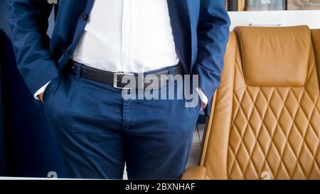 Stylish young businessman in blue suit standing in modern office with brown leather armchair Stock Photo