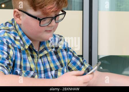 Red-haired little boy in a plaid shirt with smartphone sitting at a bar table. Cute red-haired boy with cellphone and glasses in an urban context. Stock Photo