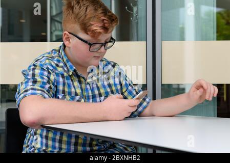 Red-haired little boy in a plaid shirt with smartphone sitting at a bar table. Cute red-haired boy with cellphone and glasses in an urban context. Stock Photo