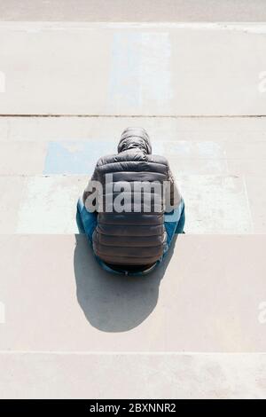 a young caucasian man, wearing jeans and a gray hooded jacket, seen from above, sitting on an outdoor concrete stairway Stock Photo