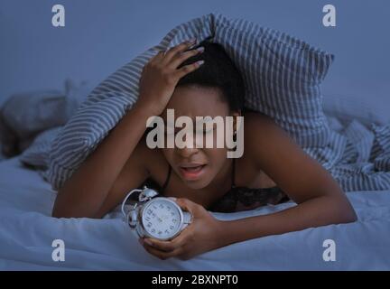 Sleepless night. Sad african american girl presses hand to head and holding alarm clock Stock Photo