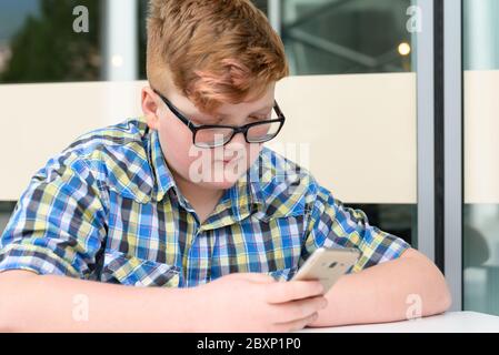 Red-haired little boy in a plaid shirt with smartphone sitting at a bar table. Cute red-haired boy with cellphone and glasses in an urban context. Stock Photo