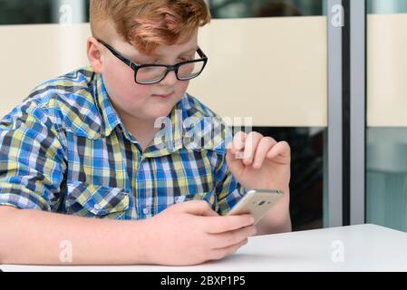 Red-haired little boy in a plaid shirt with smartphone sitting at a bar table. Cute red-haired boy with cellphone and glasses in an urban context. Stock Photo