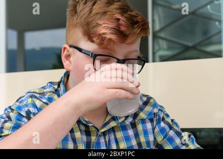 Red-haired boy with cellphone drinking from a glass of ice water with lemon. Child with glasses dressed in a plaid shirt looks at the smartphone and d Stock Photo