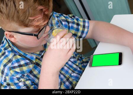 Plump red-haired boy with cellphone drinking from a glass of ice water with lemon. Child with glasses dressed in a plaid shirt looks at the smartphone Stock Photo
