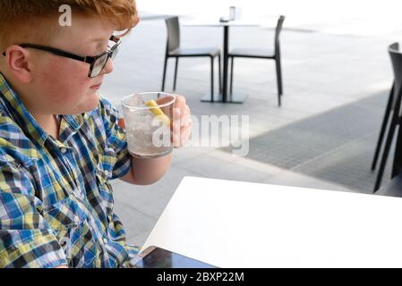 Red-haired boy with cellphone drinking from a glass of ice water with lemon. Child with glasses dressed in a plaid shirt looks at the smartphone and d Stock Photo