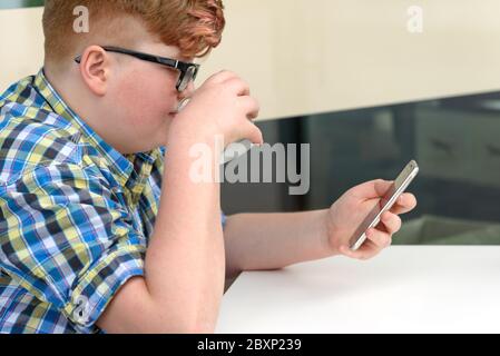 Red-haired boy with cellphone drinking from a glass of ice water with lemon. Child with glasses dressed in a plaid shirt looks at the smartphone and d Stock Photo