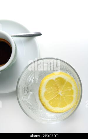 Glass of sparkling water with lemon and cup of coffee. Iced mineral water with lemon and cup of coffee on the white table. Stock Photo