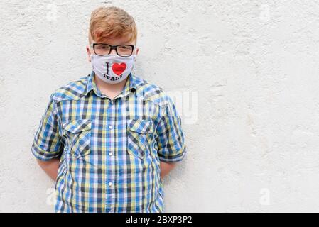 Red-haired boy with glasses and surgical mask with the inscription I LOVE ITALY. A boy in a plaid shirt stands on a wall background. Stock Photo