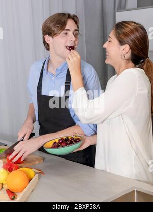 Smiling young couple at the kitchen having fun with cherries while cutting vegetables. Cooking at home and fun concept. Stock Photo