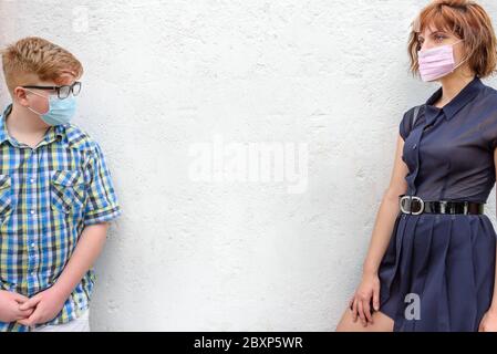 Young red-haired woman and boy wearing a surgical mask stand against a wall background. Sister with trendy glasses and little brother with glasses wea Stock Photo