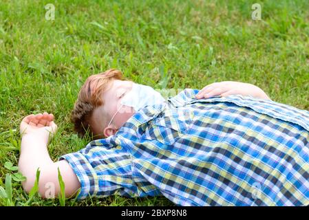 Young boy with surgical mask lying on the grass in the park near a sitting girl who smokes. Child dressed in a plaid shirt lying on the green field ne Stock Photo