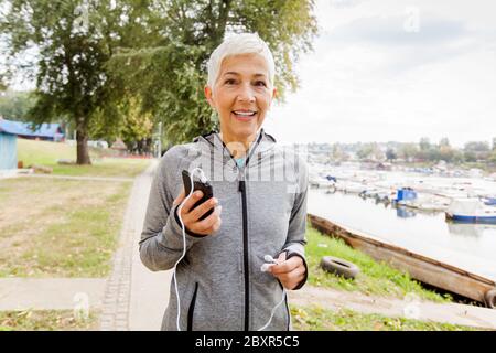 Portrait Of Active Senior Running Woman Listening Music With Smartphone In Nature, Workout Outdoor Stock Photo
