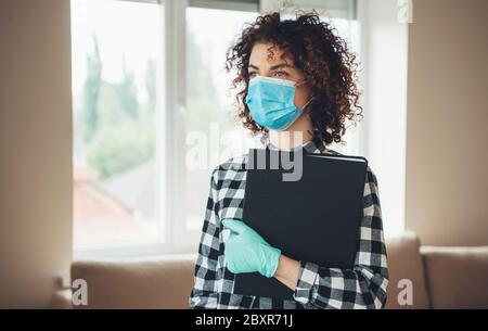 Curly haired lady with medical mask on face and gloves holding some folders is posing at home Stock Photo
