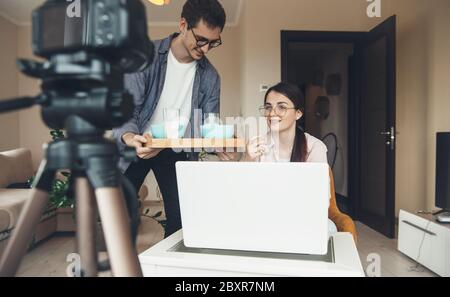 Brunette teacher with eyeglasses having an online lesson while her husband brings her breakfast Stock Photo
