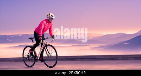 Young Woman Cyclist Riding Road Bike on the Road in the Beautiful Mountains at Purple Sunset. Adventure, Travel, Healthy Lifestyle and Sport Concept. Stock Photo