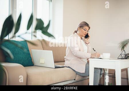 Senior caucasian woman with blonde hair and eyeglasses working from home at the pc while write something Stock Photo