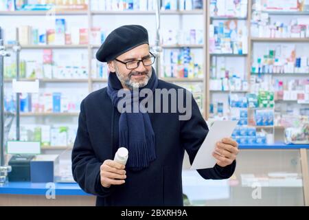 Front view of smiling eldery bearded customer wearing scarf, beret and eyeglasses standing in drugstore and choosing medcine. Man holding pills bottle in hand and reading prescription paper. Stock Photo