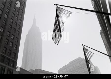 Black and white art photo from New York City with US flags hanging from buildings backlit from sun reflections and Empire State building gray in the d Stock Photo