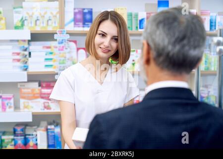 Front view of cheerful pretty woman wearing white lab coat working in chemists store with male customer. Back view of unrecognizable mature man consulting with smiling professional pharmacist. Stock Photo