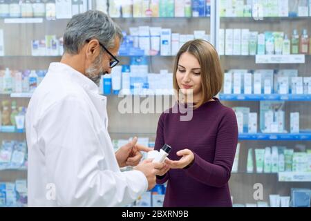 Pharmacist offering pretty female customer medical products in drugstore. Young woman holding white bottles, choosing product. Side view of mature chemist wearing glasses and white lab coat. Stock Photo
