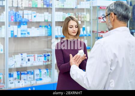 Pharmacist offering female customer medical products in drugstore. Beautiful woman holding white cosmetic bottles, choosing. Back view of unrecognizable chemist wearing glasses and white lab coat. Stock Photo