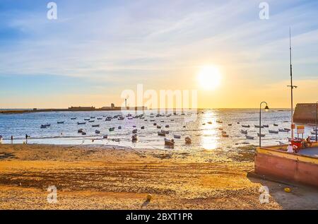 The sunset seascape with many fishing boats, moored at La Caleta beach with a view on San Sebastian Castle on the horizon, Cadiz, Spain Stock Photo