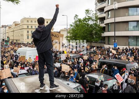 A black man stands on top of a bus stop with his fist raised above a crowd of protesters, during theBlack Lives Matters protest in London, 6 June 2020 Stock Photo
