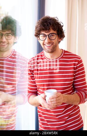 Portrait shot of happy handsome man standing by the window and having coffee while relaxing at home. Stock Photo