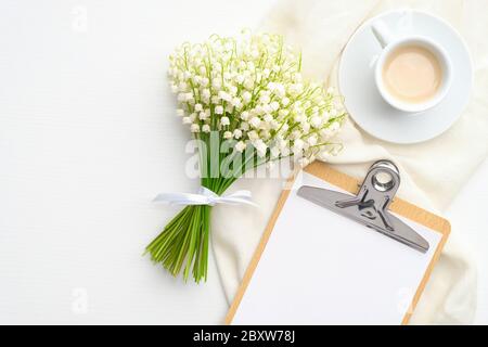 Flat lay composition with paper clipboard with blank paper list, spring flowers lily of the valley, morning coffee mug and cloth on white table. Top v Stock Photo