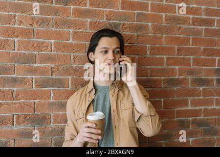 Man using mobile phone, holding cup of coffee in cafe. Hipster guy wearing stylish casual clothing holding smartphone, talking near wall. Coffee break Stock Photo