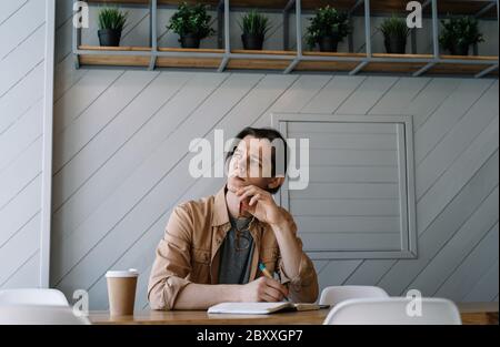 Serious man working freelance project, writing notes, thinking, brainstorming at workplace. Portrait of pensive student studying, learning language Stock Photo