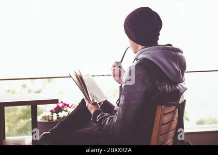 Back view on young black haired man witn piercing reeding a book and smoking tabacco pipe in outdoor cafe Stock Photo