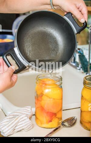 Pouring hot syrup mixture (sugar and water mixture) into a jar of peach halves until the peaches are covered,  leaving exactly 1/2 inch of space at th Stock Photo