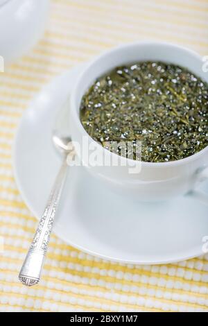White ceramic cup of Sen-cha green tea, that has freshly poured dried green tea leaves still floating on the top and seeping, and a spoon on the sauce Stock Photo