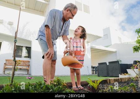 Smiling grandfather and granddaughter in backyard garden together, holding watering can have fun plants. Two generation fun time outdoor activity Stock Photo